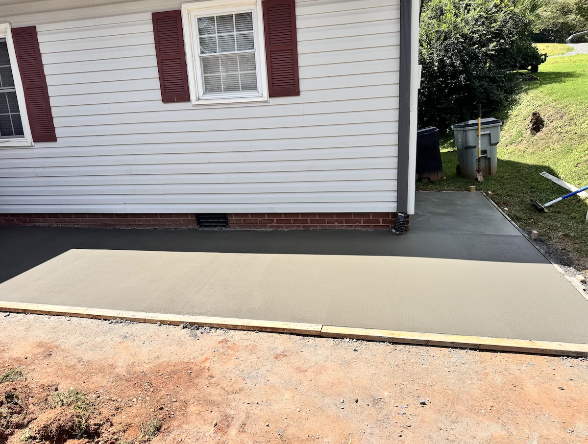 New concrete parking pad curing inside timber edge forms alongside a brick-skirted home in the NC Foothills &mdash; The Foothills Concrete Company
